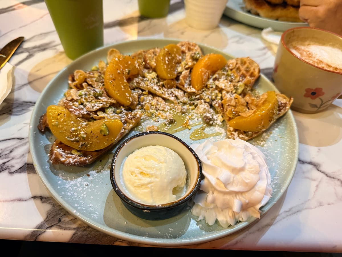A plate of Norwegian waffles with sliced peaches, ice cream, and whipped cream, plus pistachios and honey drizzle, with cappuccino and other drinks on the table.