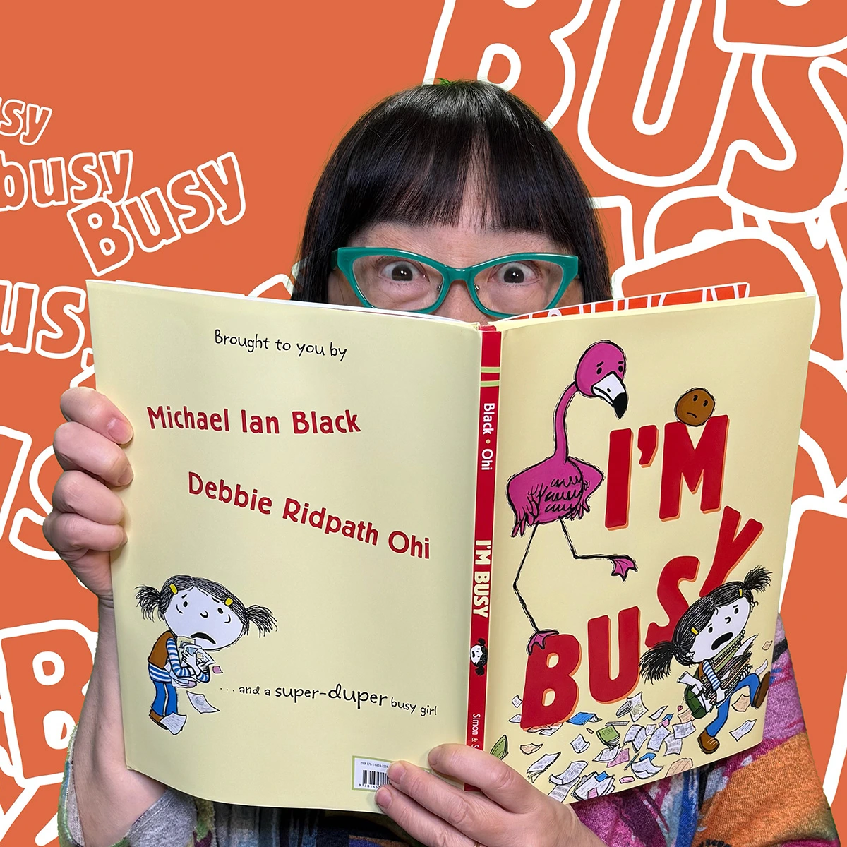 Person peeks over an open copy of the picture book I'm Busy, with only their eyes and teal glasses visible above the book. The front and back covers are visible, showing the title, author Michael Ian Black, illustrator Debbie Ridpath Ohi, a pink flamingo, and a busy child. The background is orange with repeated white outlines of the word “BUSY.”