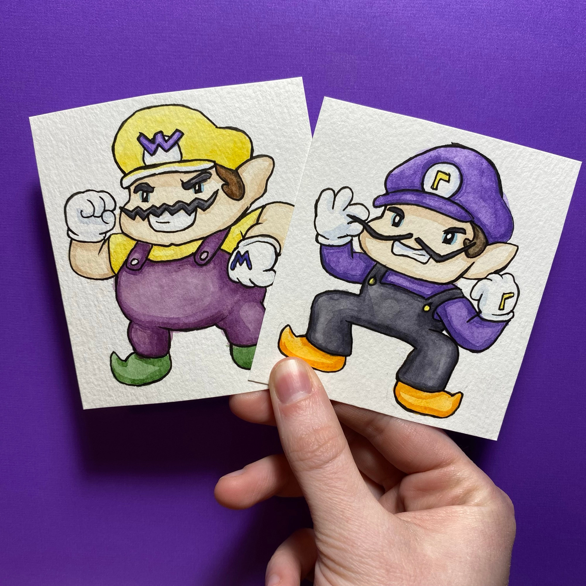 Hand holding two small paintings, one with a caricature of Wario and one is a caricature of Waluigi from Nintendo games.