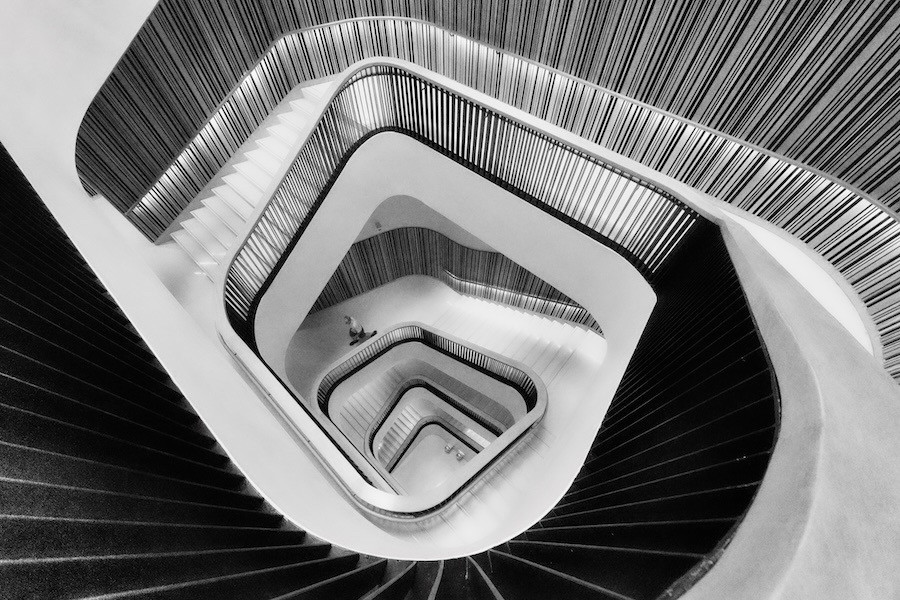 Photograph of a staircase in black and white, viewed from above, spiraling down in a square shape. A person walks down them as seen from above.