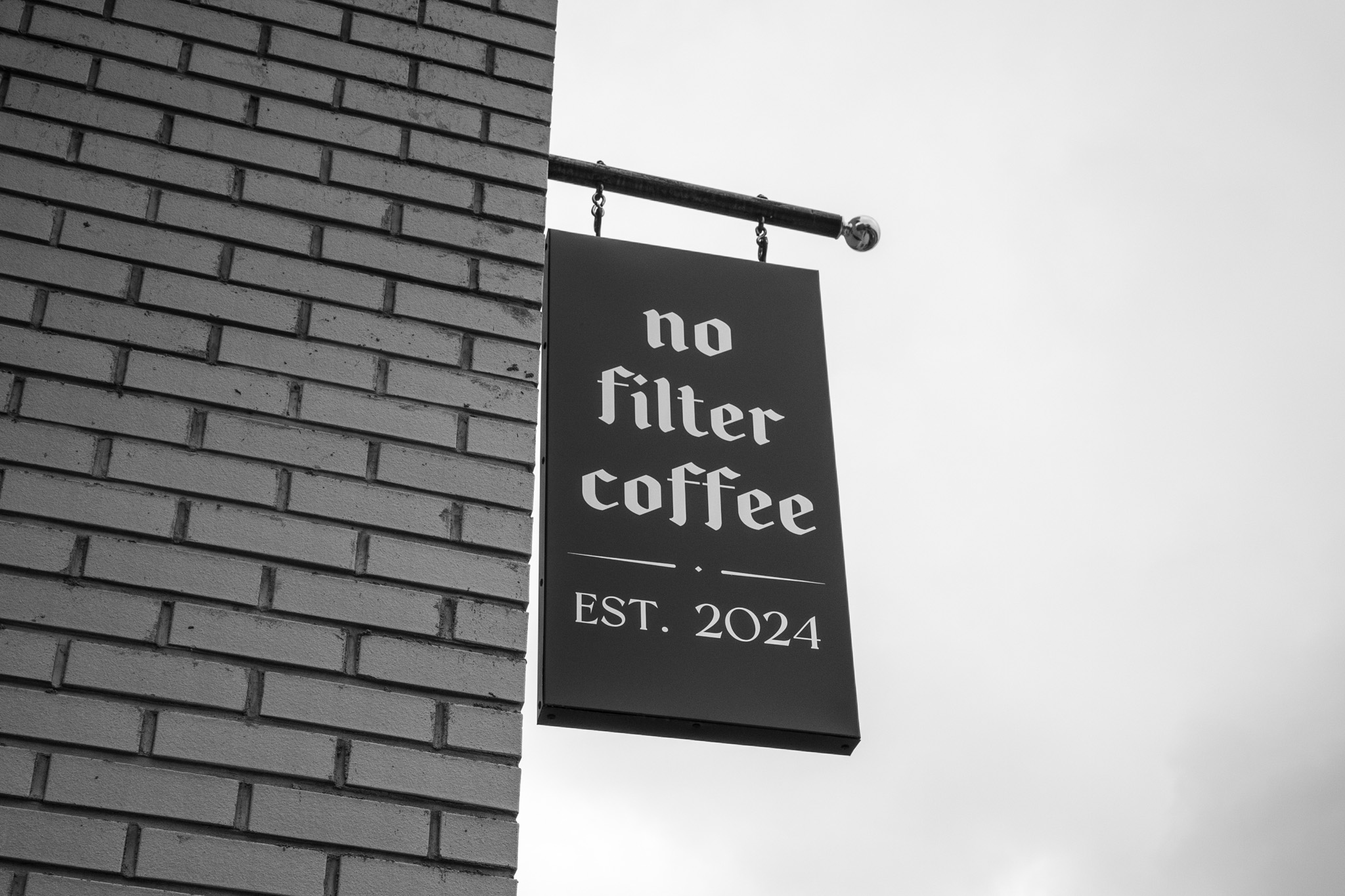 Photograph of a black sign reading "no filter coffee EST. 2024" hanging from a brick wall.