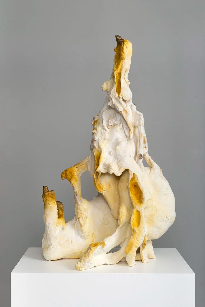 Photograph of an abstract sculpture in white and yellow, displayed on a white pedestal against a gray background. The sculpture has an organic, textured form, with a tall, slender section rising from a broader base. Yellow accents highlight the contours and crevices of the piece.