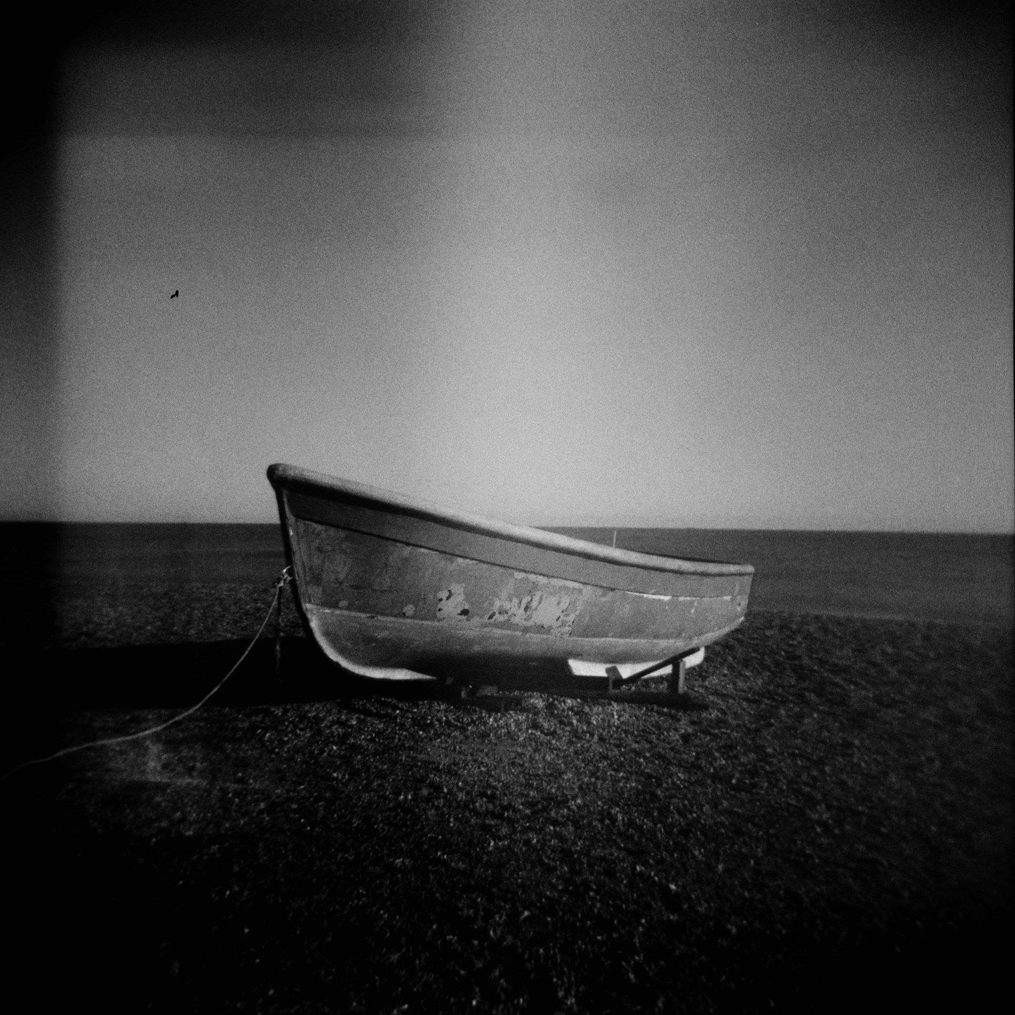 Black and white Holga photo of a lone fishing boat with its bow towards the left of the photo, beached on the shingle, with the sea and a clear sky behind