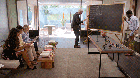 several people in a modern living room. an older man in suit and bowtie is apparently again writing on a blackboard: people = good