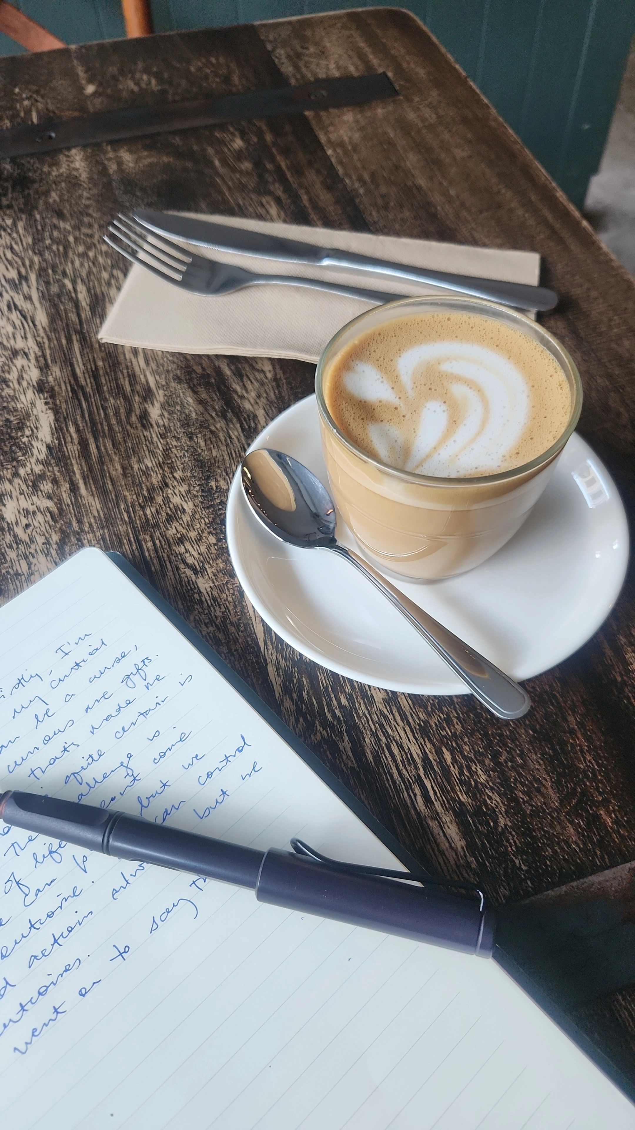  A journal sits on a cafe table. We see a fountain pen resting on the page. There's a coffee in the background.