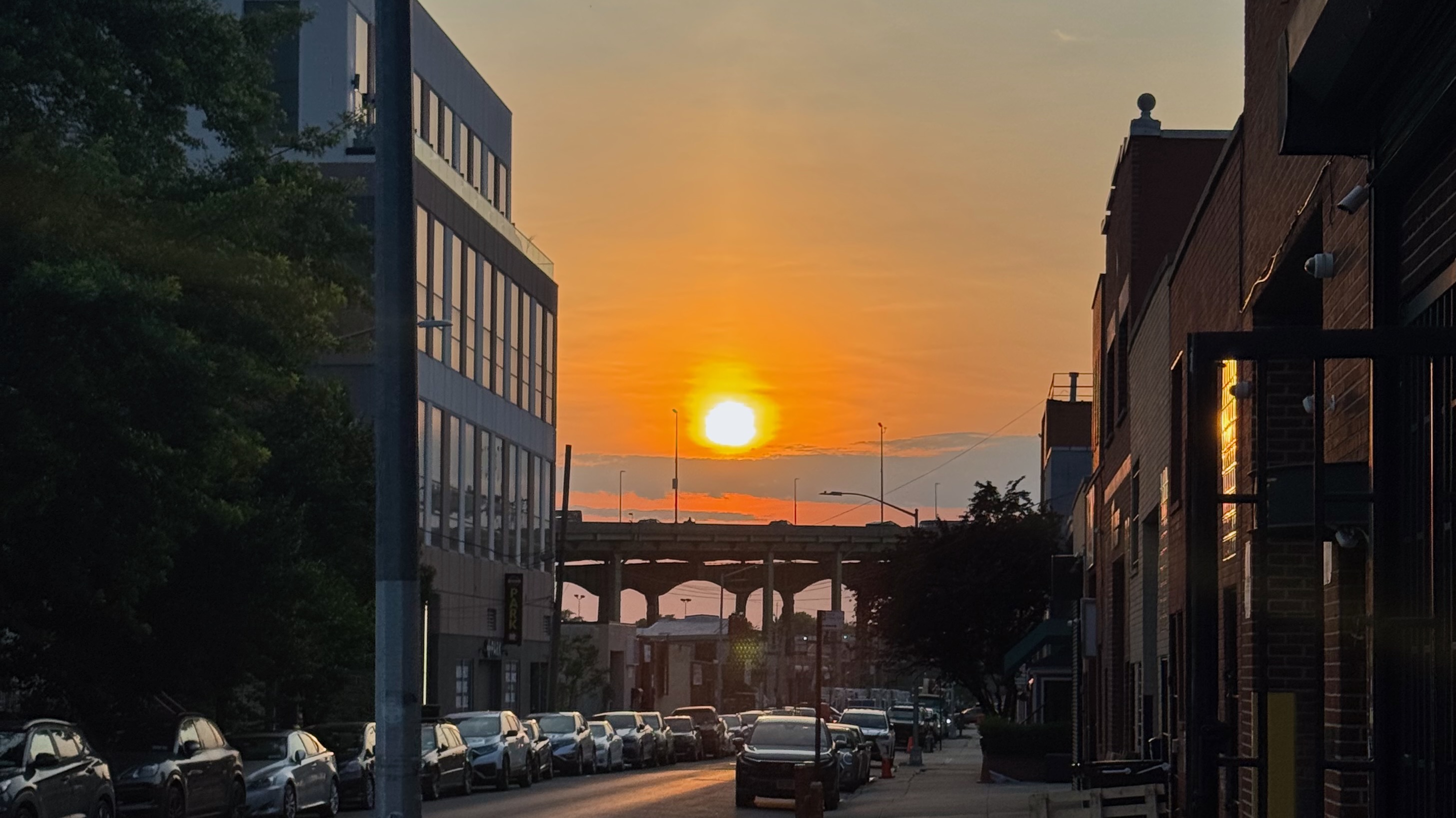 Photo of a sunset over the BQE, in Gowanus, Brooklyn, looking down a mixed use block with buildings on either side, sunlight reflecting off windows and cars.