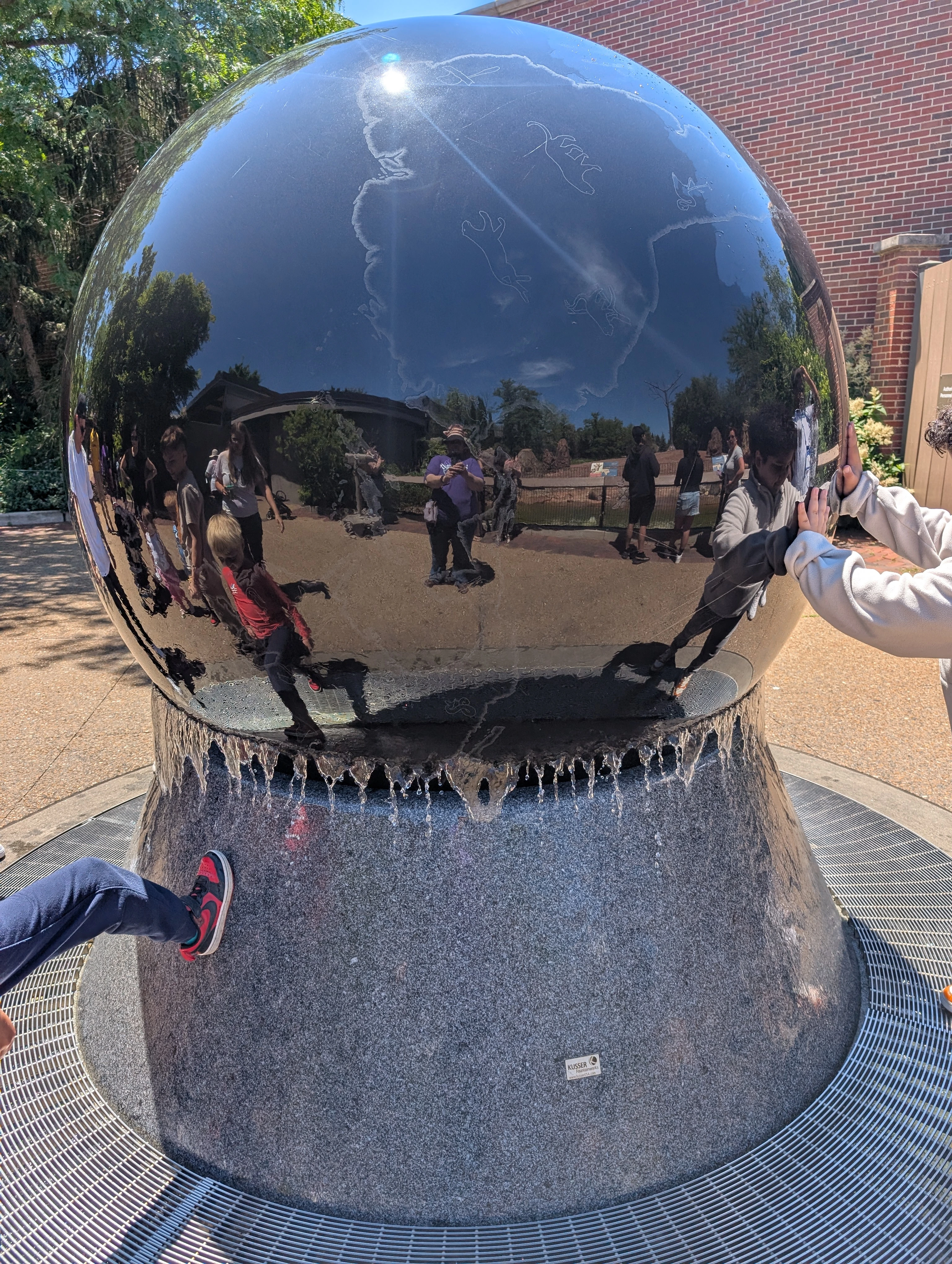 A large, black globe on a granite pedastel. You can partially see a map of Africa engraved on it, with engravings of a couple of large feline creatures. Children are touching the fountain, one of whom has his foot propped on the pedestal. There are many people reflected in the globe's surface.