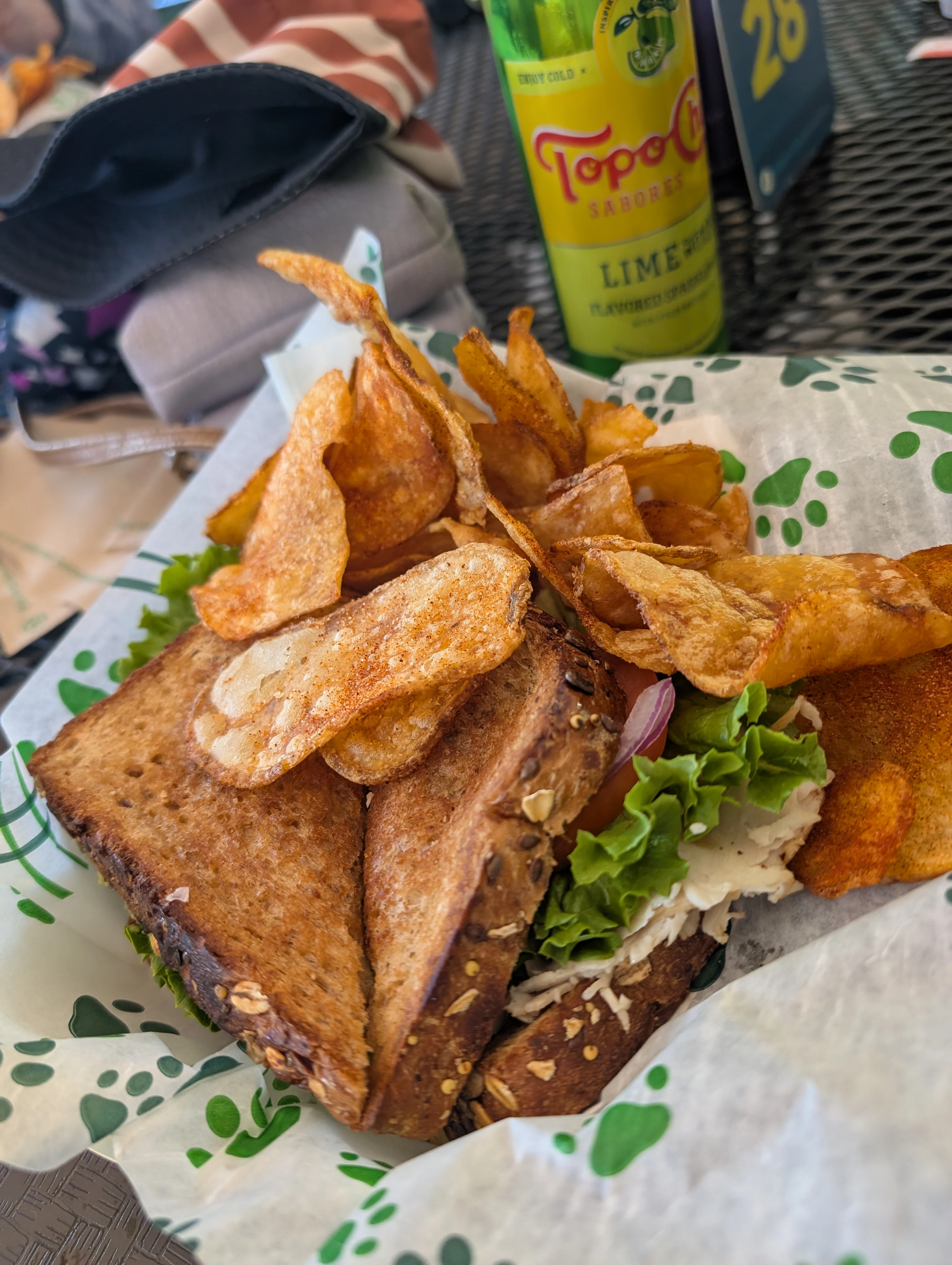 A smoked turkey sandwich with lettuce, onion, tomato, and avocado on a whole-grain bread. Piled on top are thick-cut BBQ-seasoned potato chips. There's a can of mint-lime Topo-Choco sparkling water in the background.