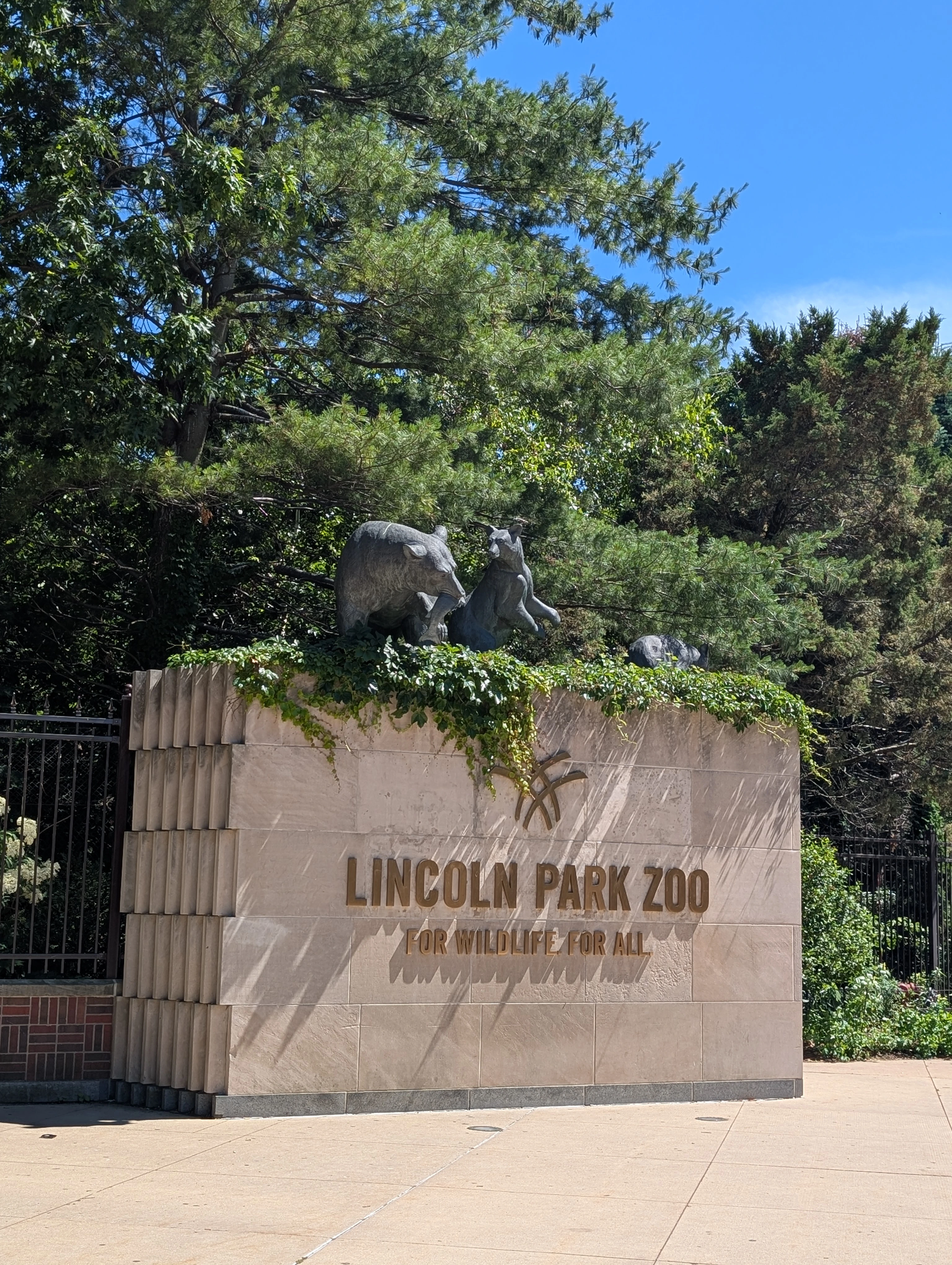 A stone pedastal with metal letters attached. It says Lincoln Park Zoo: For Wildlife, For All. There is a bronze bear family cavorting on top in the greenery.
