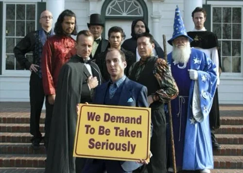 A promotional group photograph from the comedy series *Arrested Development* features the fictional organization "The Alliance of Magicians" assembled on brick steps outside a white building. In the front center, a deadpan Gob Bluth (played by Will Arnett) wears a dark blue suit while holding a prominent yellow sign that reads, **"We Demand To Be Taken Seriously."** He is flanked by nine other men in eclectic, theatrical magician attire, including a man in a black cape, another holding a wooden staff, and an older man dressed in a bright blue wizard robe and star-patterned pointed hat. Posing with hands on hips in the upper-left background is a bald man with glasses—who is frequently mistaken for Jim Rash (Dean Pelton from *Community*) but is actually show director Greg Mottola making a cameo.