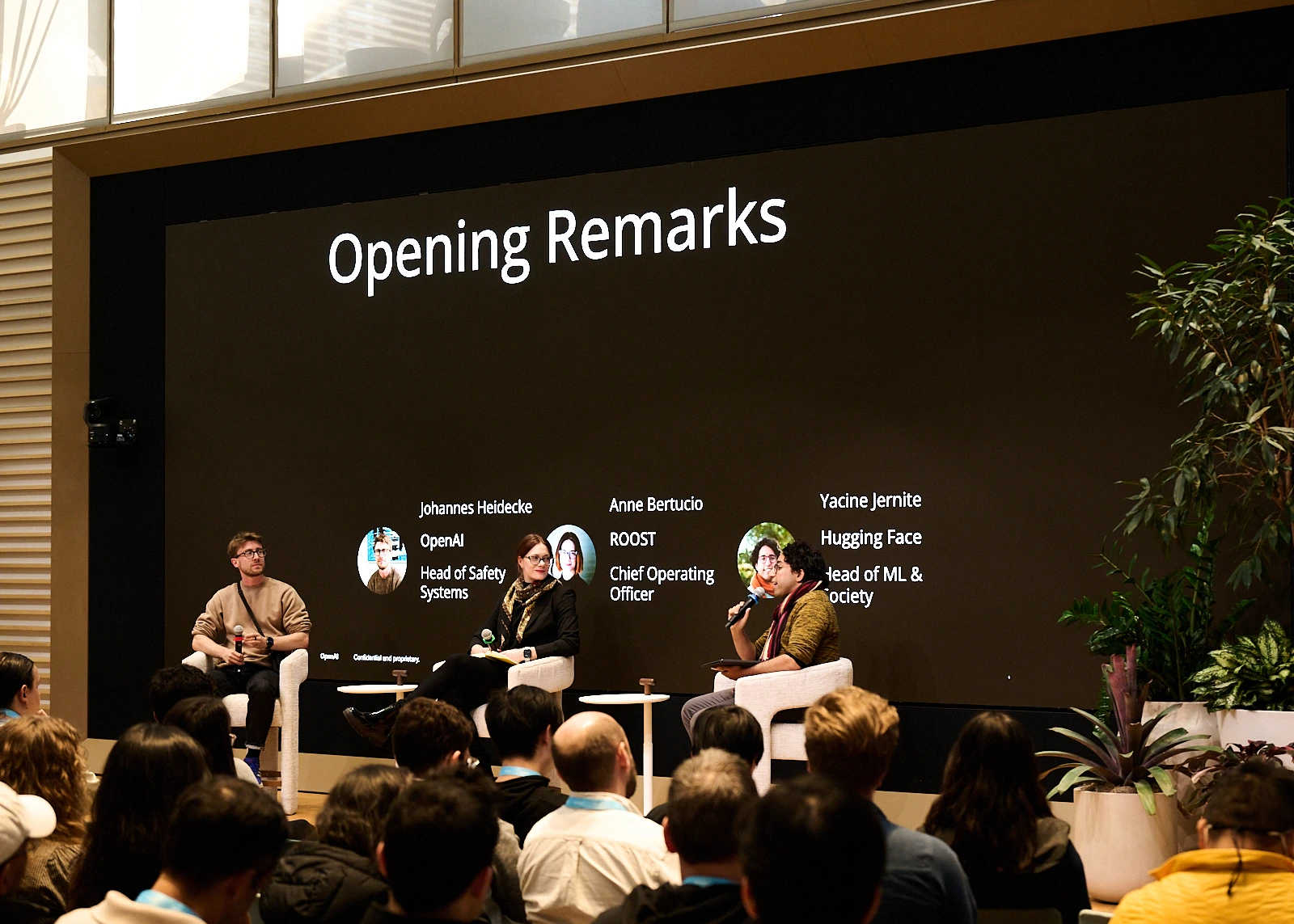 three people sit on stage with a large screen behind them that says "Opening Remarks": Johannes Heidecke, head of Safety Systems at OpenAI; Anne Bertucio, COO at ROOST; Yacine Jernite, Head of ML & Society at HUgging Face