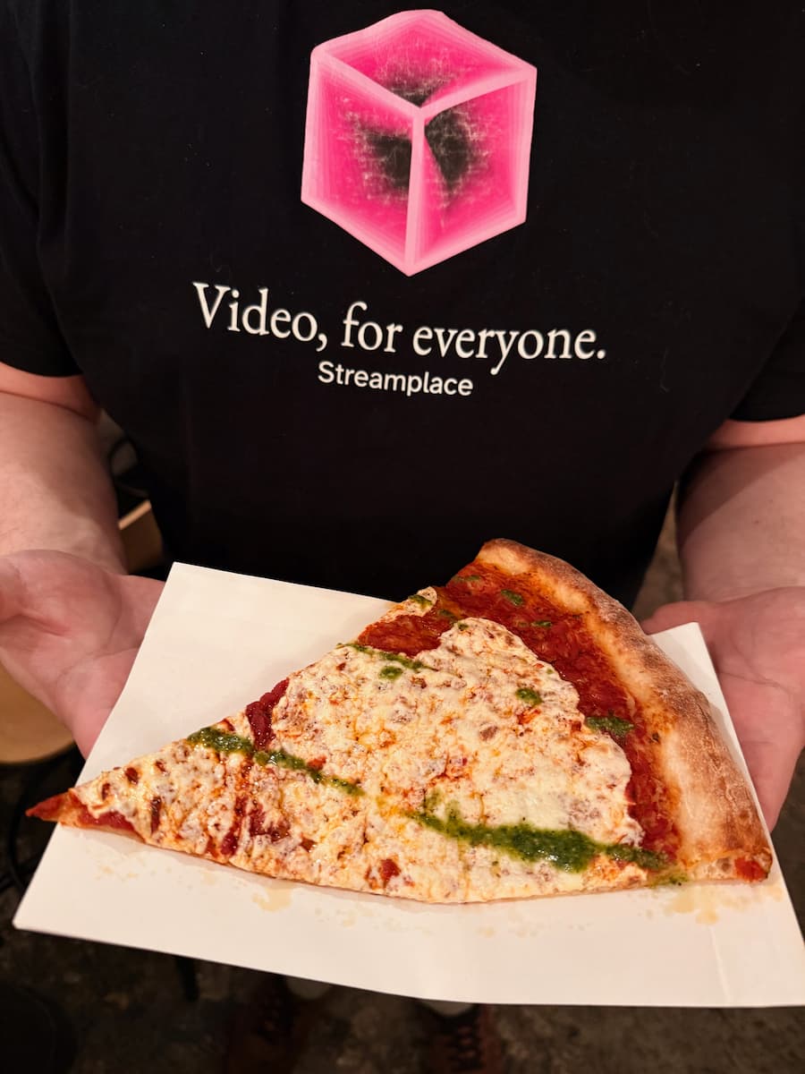 A man wearing a t-shirt with a pink cube and the slogan "Video, for everyone. Streamplace". The man is holding pizza, because also everyone should have pizza.
