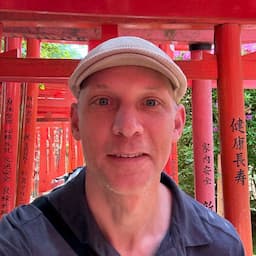 Jim in front of a Japanese style shrine made of red pillars and beams. He is wearing a cap.