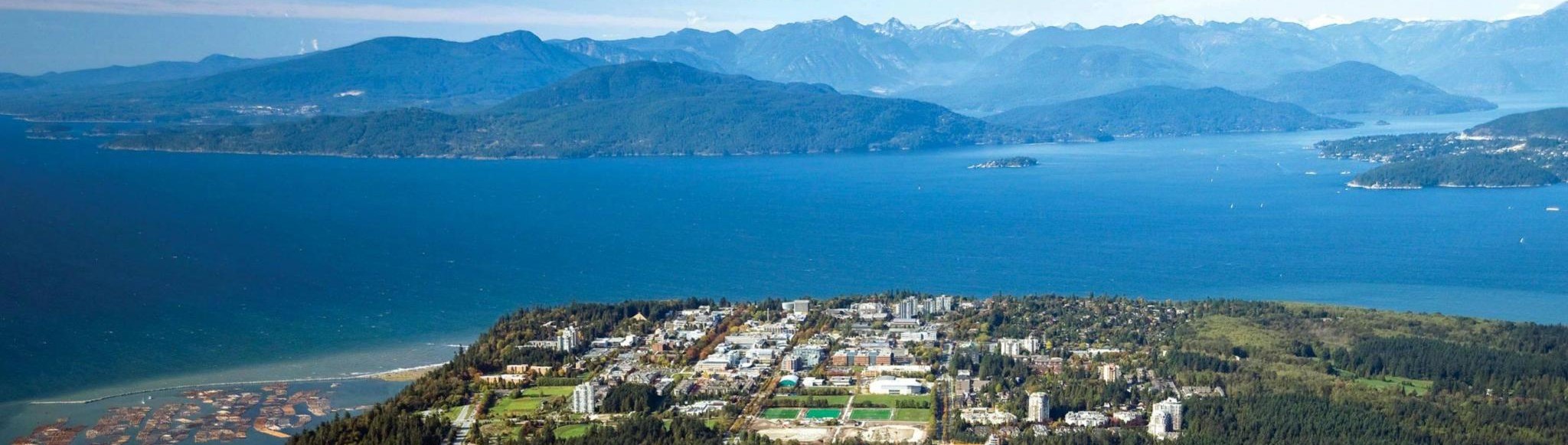 Aerial photograph of The University of British Columbia. A number of buildings are encapsulated by evergreens in the foreground, and a water inlet with hills and mountains encapsulate it in the background.
