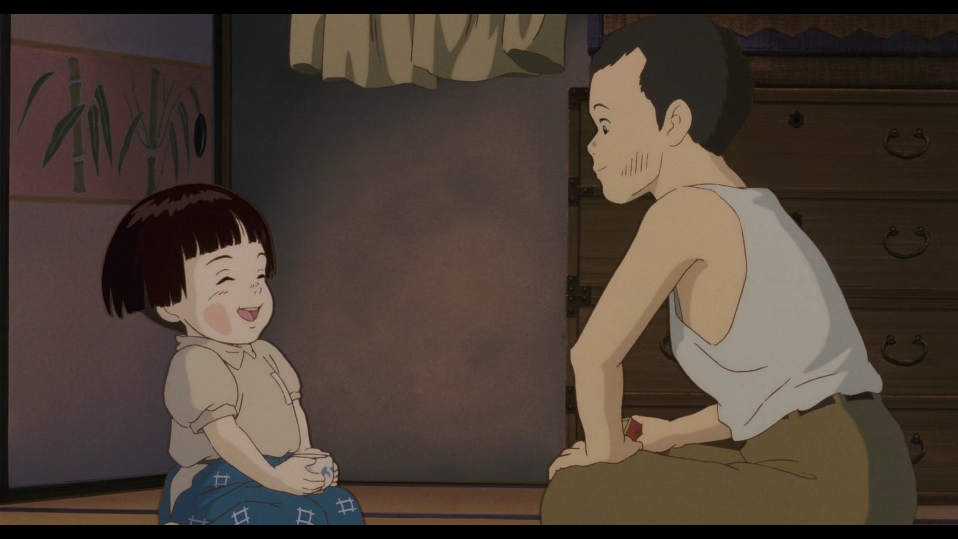 Scène un peu tendre du Tombeau des Lucioles
A gauche, Setsuko, petite fille japonaise de 4 ans, les cheveux noirs, courts, coupés en frange au front, portant un haut gris, et un pantalon bleu avec des carreaux blancs, tenant une tasse en porcelaine est assise à genoux sur un parquet. Elle a les yeux fermés et semble rire à gorge déployée.
Face à elle, à droite de l'image, se tient Seita, adolescent japonais de 14 ans, cheveux noirs, yeux noirs, vêtu d'un débardeur blanc et d'un pantalon marron, assis en tailleur, qui regarde sa sœur avec tendresse. On devine qu'il tient un objet rouge et jaune à la main [une boîte en fer].
En arrière plan, les murs blancs de leur chambre, aves à droite une esquisse de bambou qui décore la pièce, un rideau jaune en haut et une commode à droite.