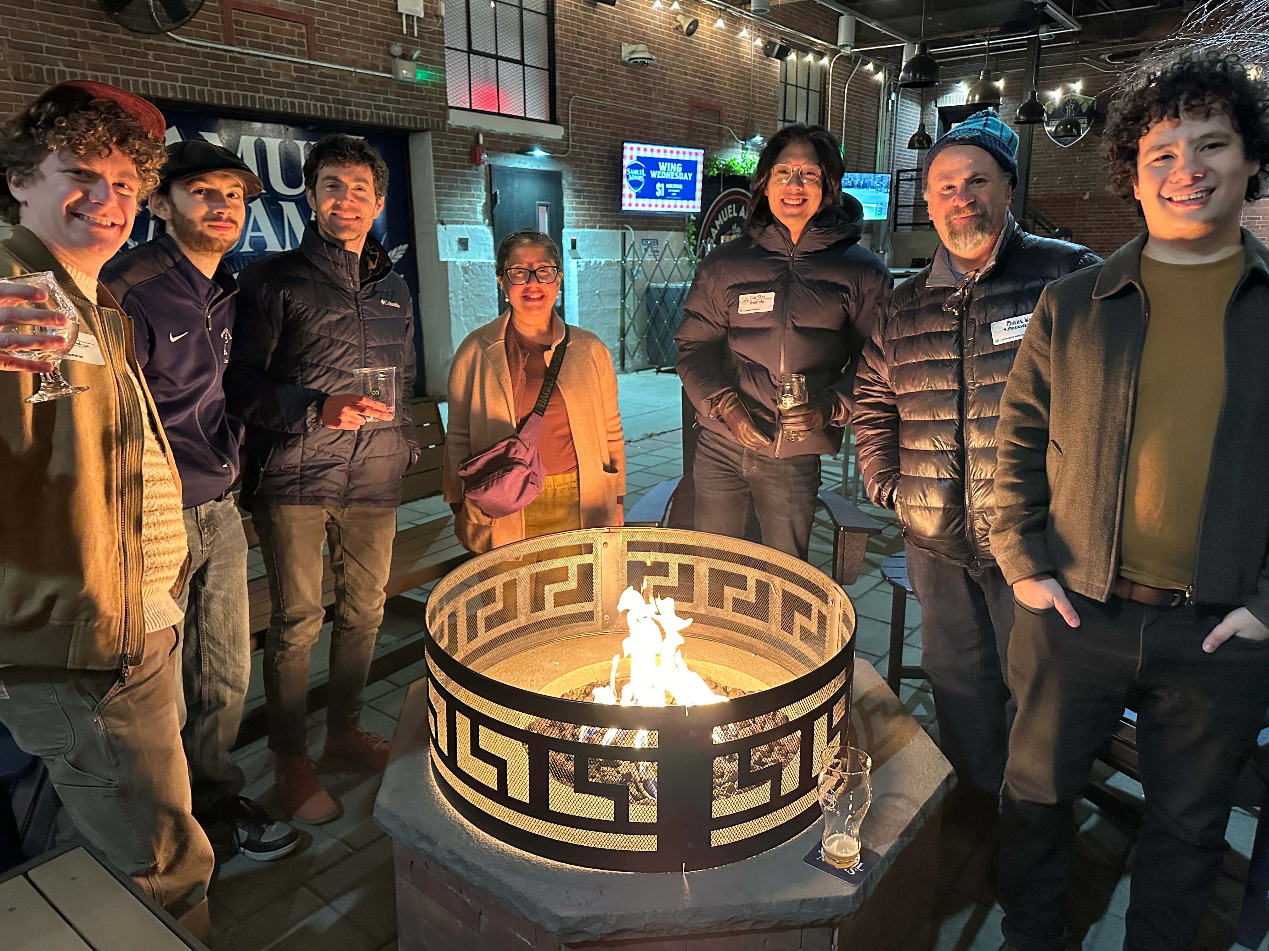 A group of people standing around a fire pit in front of Sam Adams Boston Brewery.