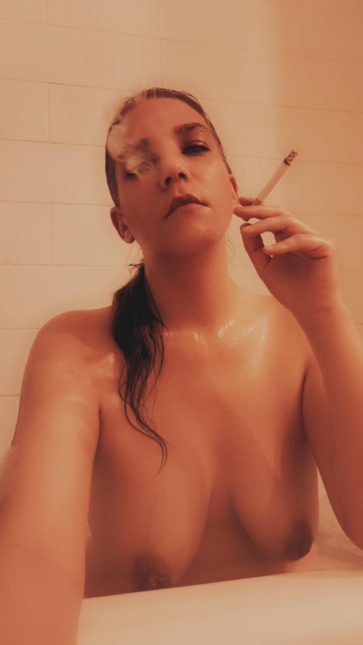 Valerie Rayne exhales a cork-filtered cigarette while her brown hair is in a ponytail swept over her shoulder.
