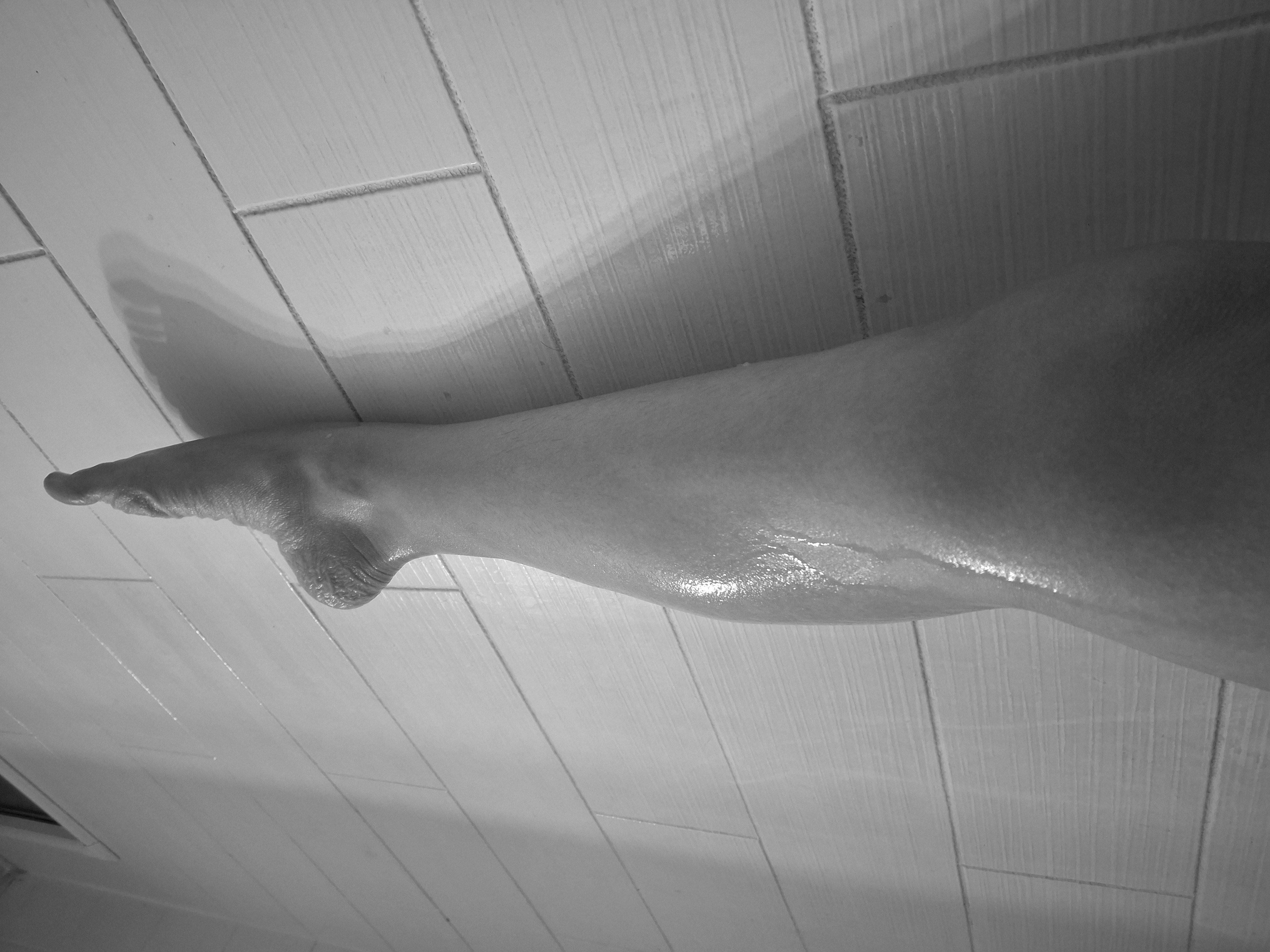 Valerie Rayne rests her leg on the bathroom wall while pointing her wrinkled foot in this monochrome photo.