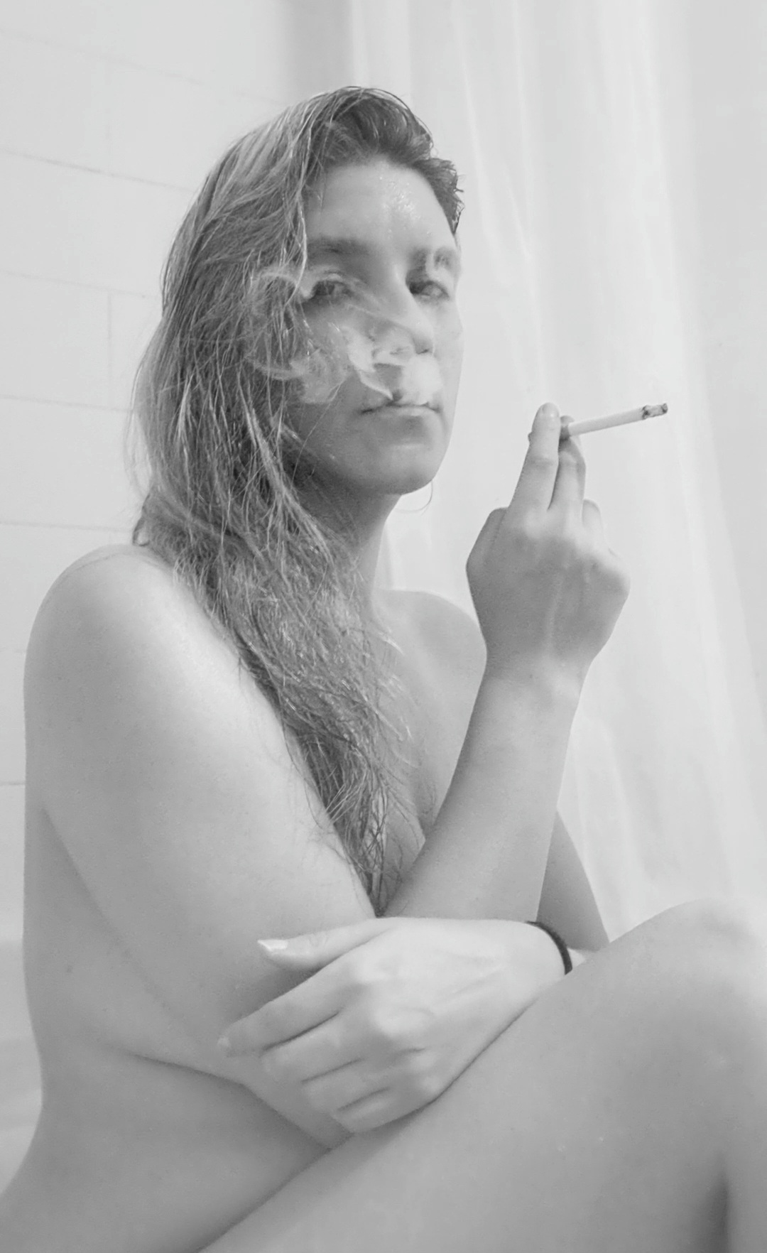 Valerie Rayne, in black and white, sitting in the bath exhaling a tendril of smoke while her long gray/brown hair is swept over one shoulder. She holds a cork-filtered cigarette between her index and middle finger while her other hand is crossed over her body.