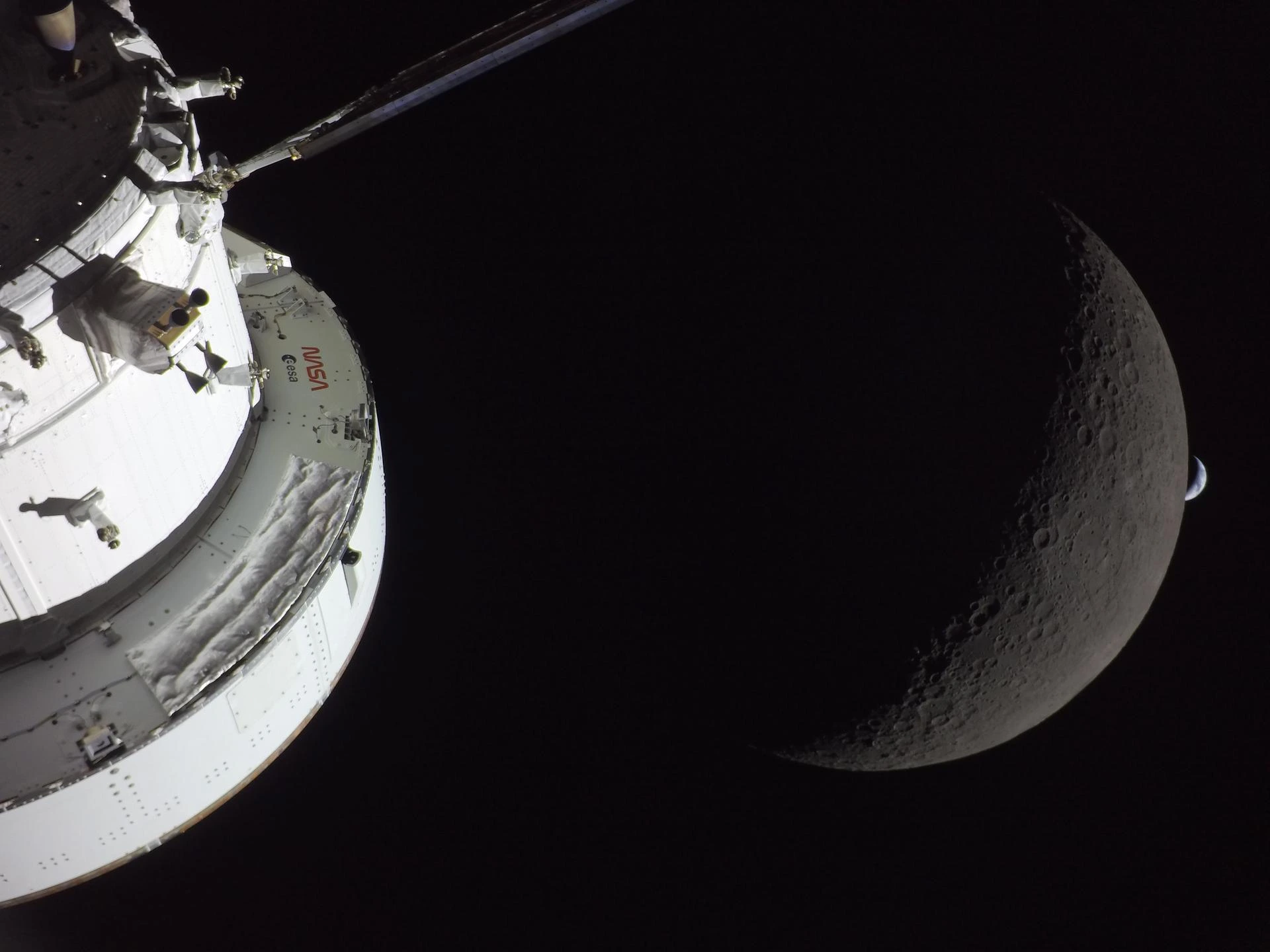 NASA.gov Bild von der Artemis II Mission, April 2026. Das weisse Orion-Raumschiff ist im Vordergrund links zu sehen. Rechts davon der Mond als beleuchtete, graue Sichel, und rechts des Mondes die blaue Sichel der weit entfernten und deshalb sehr klein erscheindenen Erde. Eingebettet sind die drei in tiefes Schwarz. 