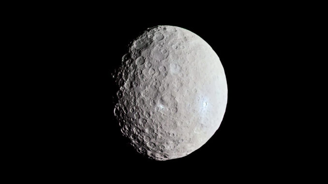 Ein Bild der Ceres von der Raumsonde Dawn. Schräg von rechts beleuchtet, steht die metallgraue, stark zerkraterte und von gelegentlichen weissen Flecken bedeckte Oberfläche von Ceres im starken Kontrast zum schwarzen Weltraum darum herum. 