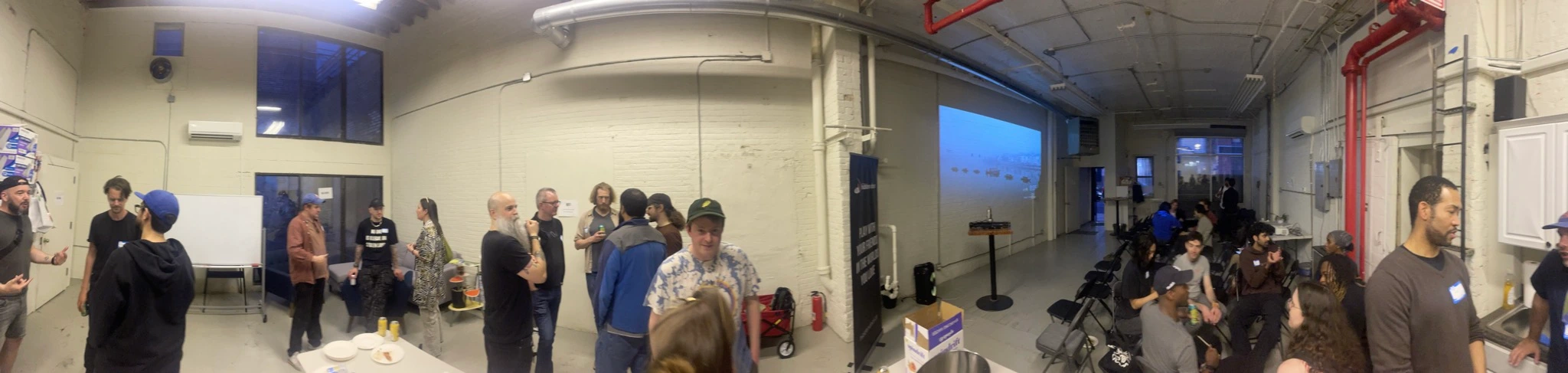 panorama photo of a large room with tables and people standing on one side, and projector screen and chairs on the other side with some people on the other side, a couple dozen people visible