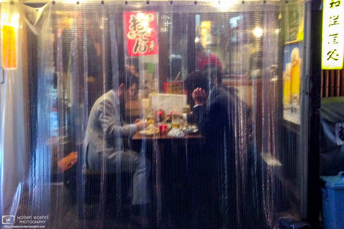Two salarymen are enjoying a simple dinner seated outside a pub in the Nakano area of Tokyo, Japan, as seen through a plastic blind outside the shop. © 2013 Norbert Woehnl Photography.