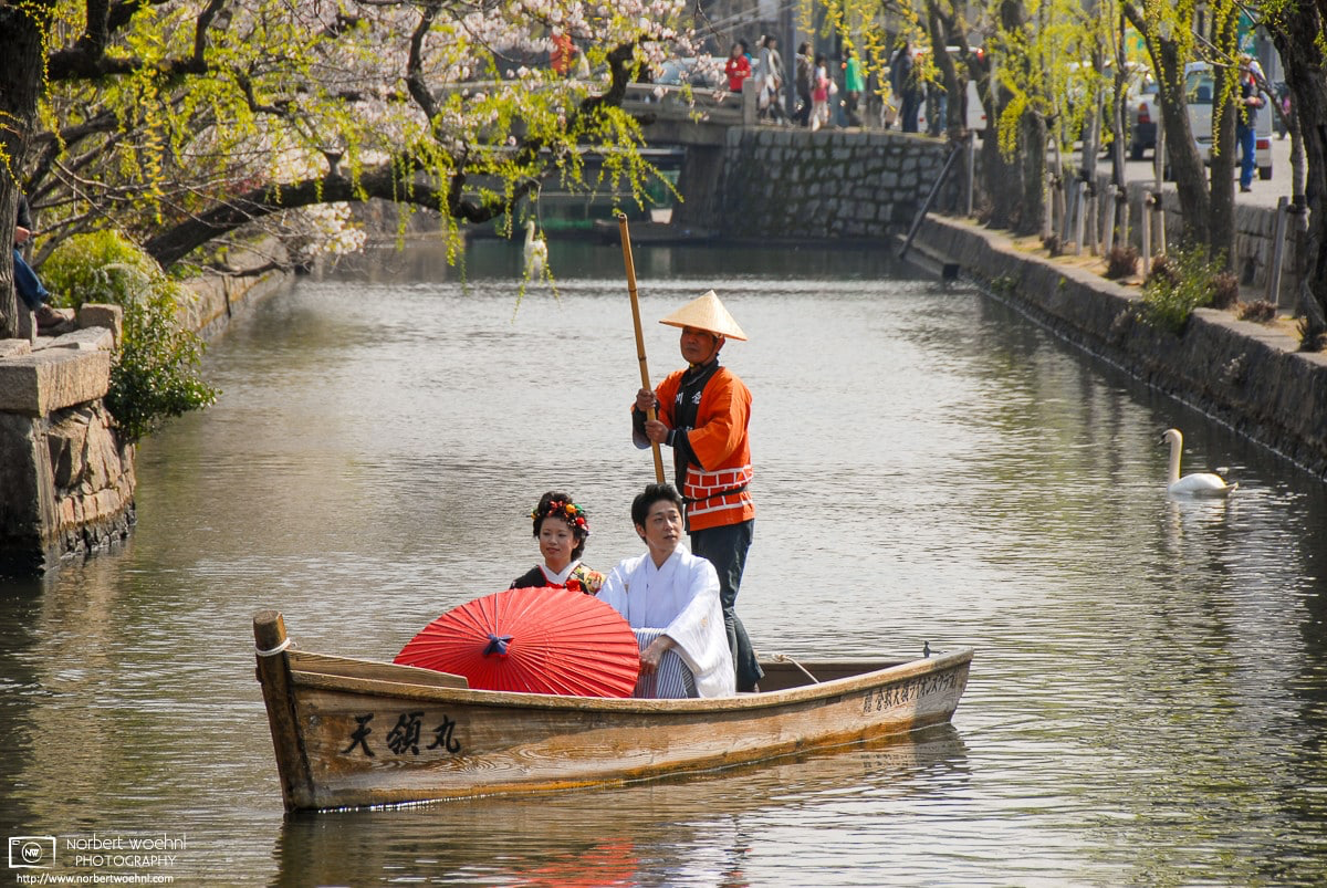 A man and a woman in traditional clothes and holding a paper umbrella are riding in a boat on the canal in the Bikan District of Kurashiki in Okayama Prefecture, Japan. © 2009 Norbert Woehnl Photography.