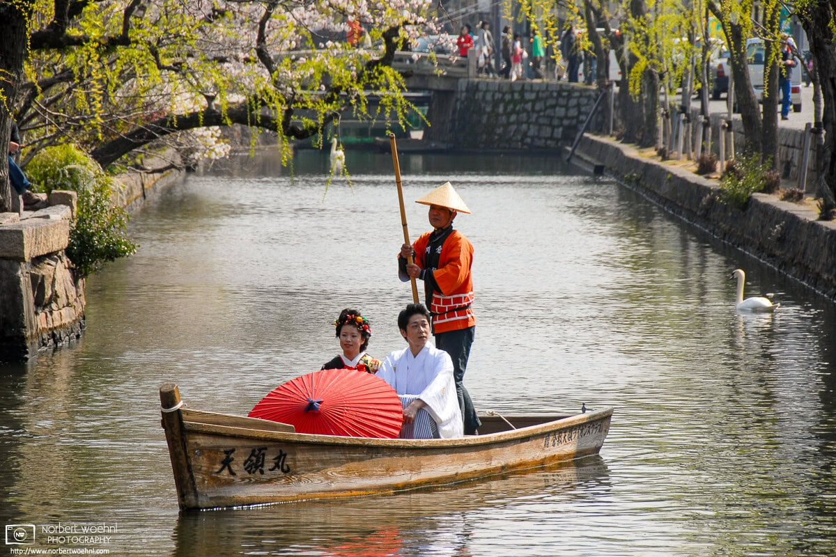 A man and a woman in traditional clothes and holding a paper umbrella are riding in a boat on the canal in the Bikan District of Kurashiki, Japan. © 2009 Norbert Woehnl Photography.