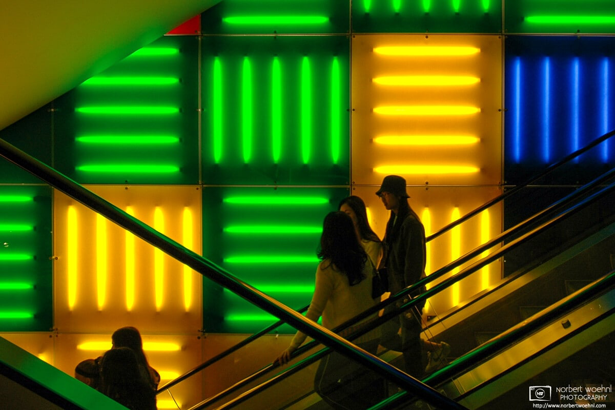 Against a striking background pattern of colorful neon lights, people are riding an escalator at a store on Times Square in Manhattan, New York. © 2005 Norbert Woehnl Photography.