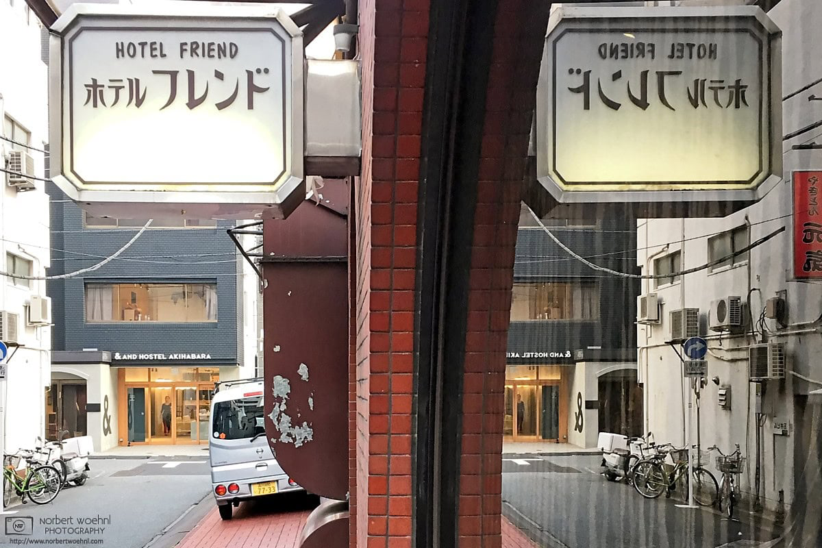 In the Taito Ward of Tokyo, Japan, a Showa-era hotel sign is reflected in a window, opening up a view towards a quiet backstreet with a more contemporary hotel. © 2018 Norbert Woehnl Photography.