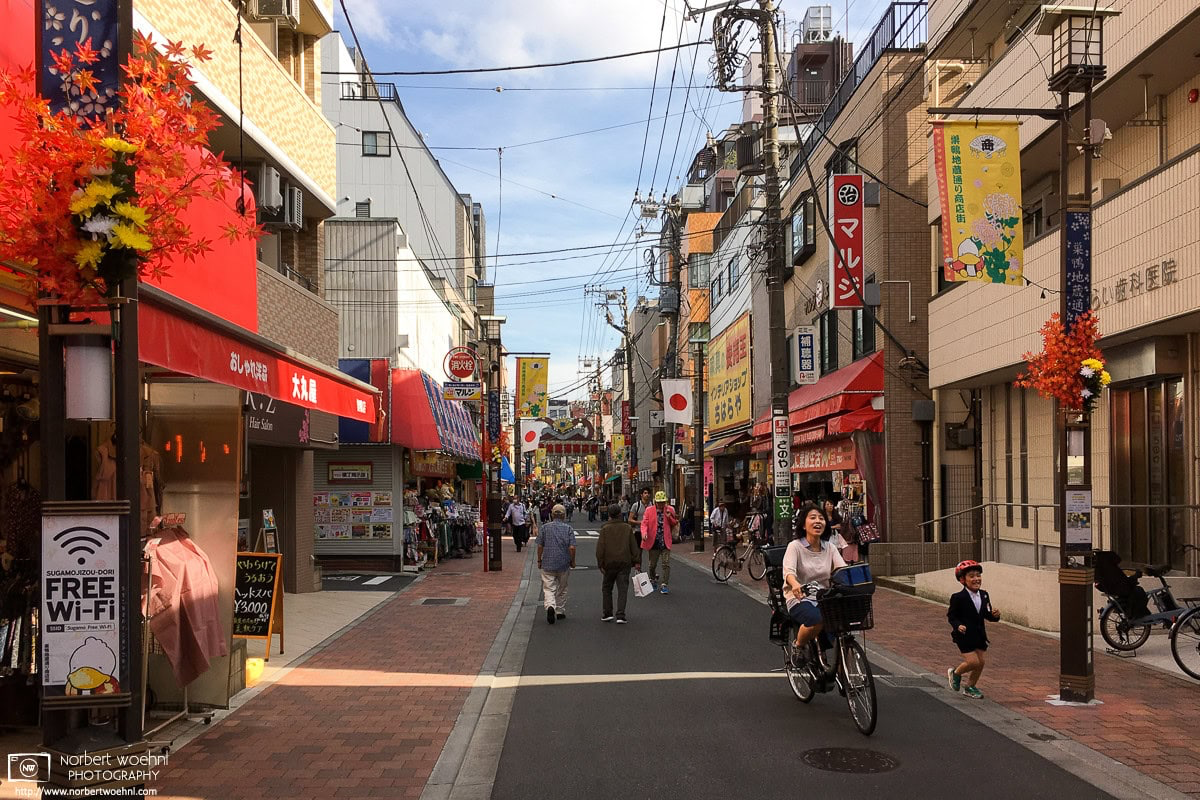 A boy is joyfully running beside his bicycling mother on Sugamo Jizodori Shopping Street in the Toshima Ward of Tokyo, Japan. © 2018 Norbert Woehnl Photography.