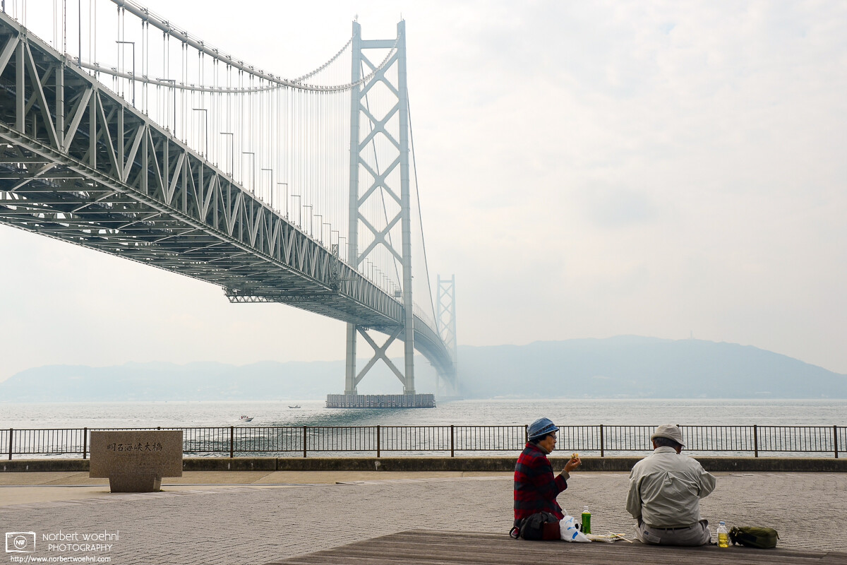 A woman and a man are having some food and drinks on a pier adjacent to Akashi Kaikyo Bridge in Kobe, Japan. © 2011 Norbert Woehnl Photography.