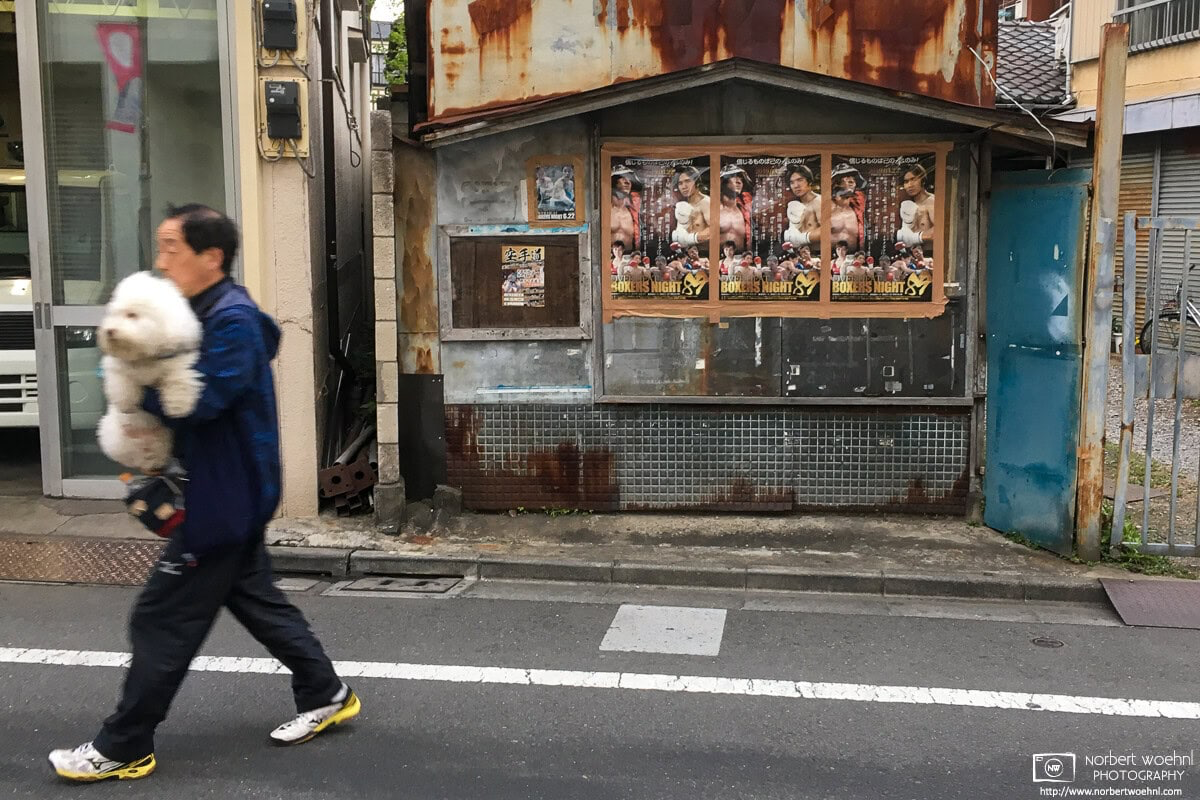 A man carries a white dog in front of an old kiosk along the border of Itabashi and Toshima Wards in Tokyo, Japan. © 2019 Norbert Woehnl Photography.
