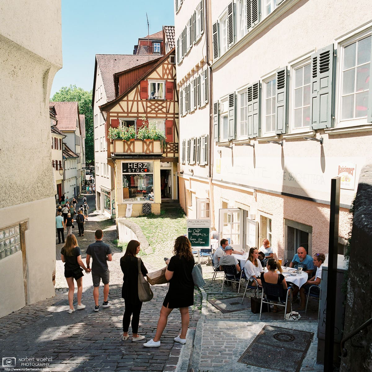 In the historic city center of Tübingen, Germany, people are seated outside a restaurant, or casually strolling between half-timbered buildings on a cobblestone street. © 2017 Norbert Woehnl Photography.