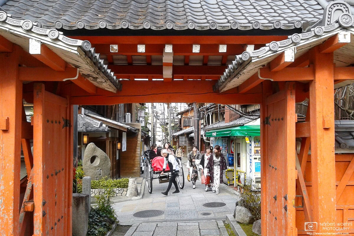 In the Higashiyama area of Kyoto, Japan, an orange temple gate frames a scene of kimono-clad women and a rickshaw driver about to make a turn. © 2012 Norbert Woehnl Photography.
