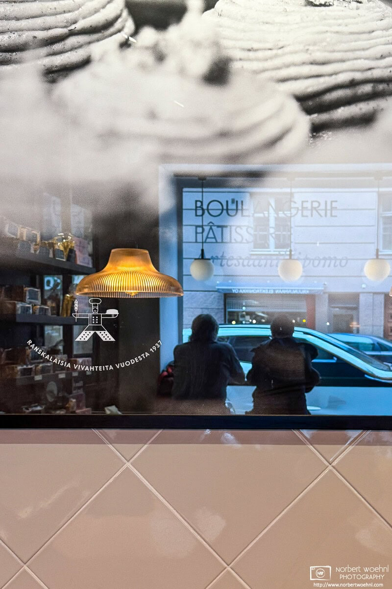 While having lunch at a café in the Kruununhaka district of Helsinki, Finland, I noticed this reflection of other customers in a framed image of pastries. © 2026 Norbert Woehnl Photography.