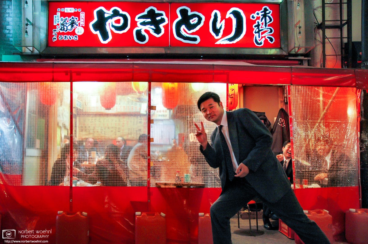 A salaryman in Tokyo, Japan, is posing for my camera outside a Yakitori stall, much to the amusement of his colleagues watching from behind. © 2006 Norbert Woehnl Photography.