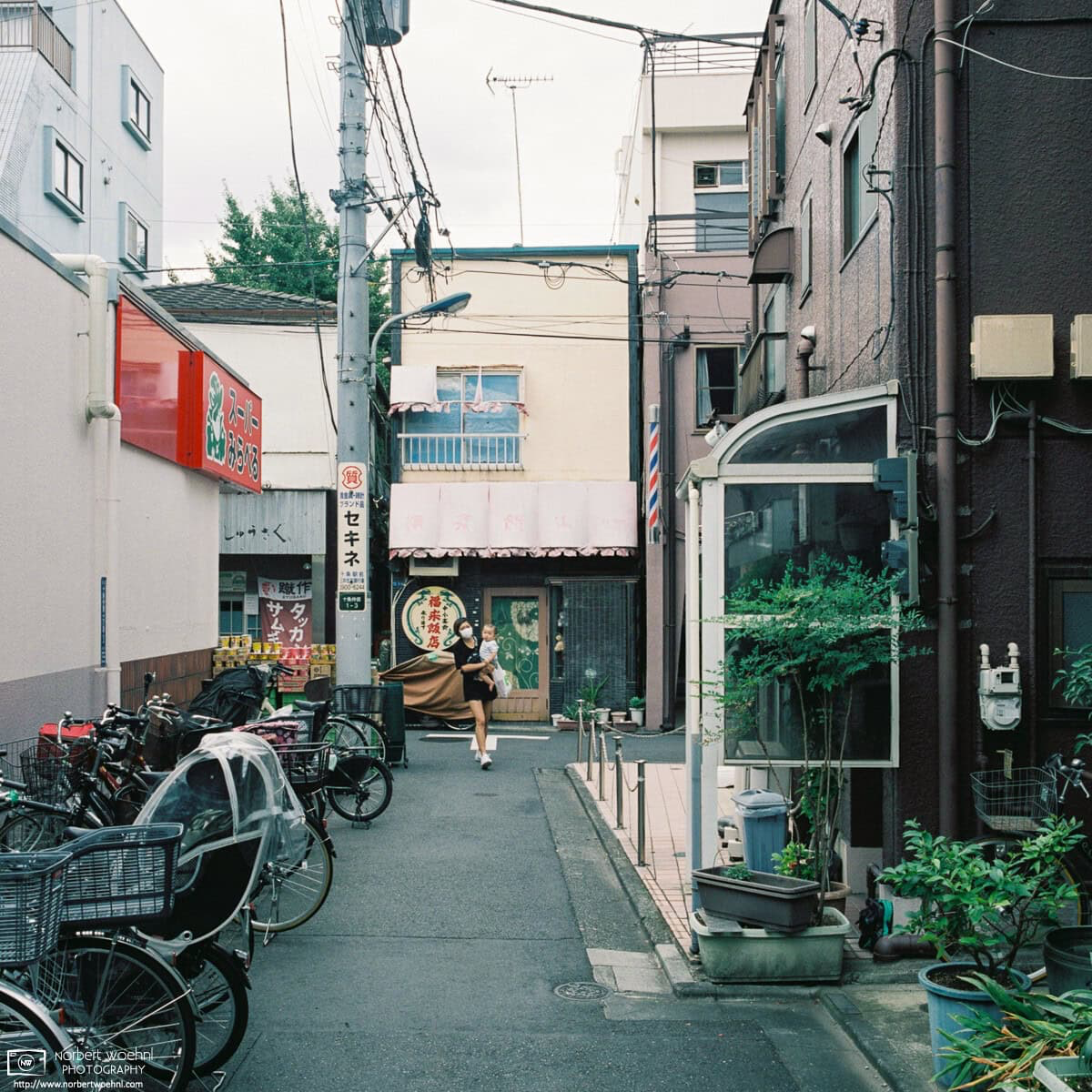 A young mother carrying her child is turning the corner at a side street supermarket in the Jujo area of Kita Ward in Tokyo, Japan. © 2021 Norbert Woehnl Photography.