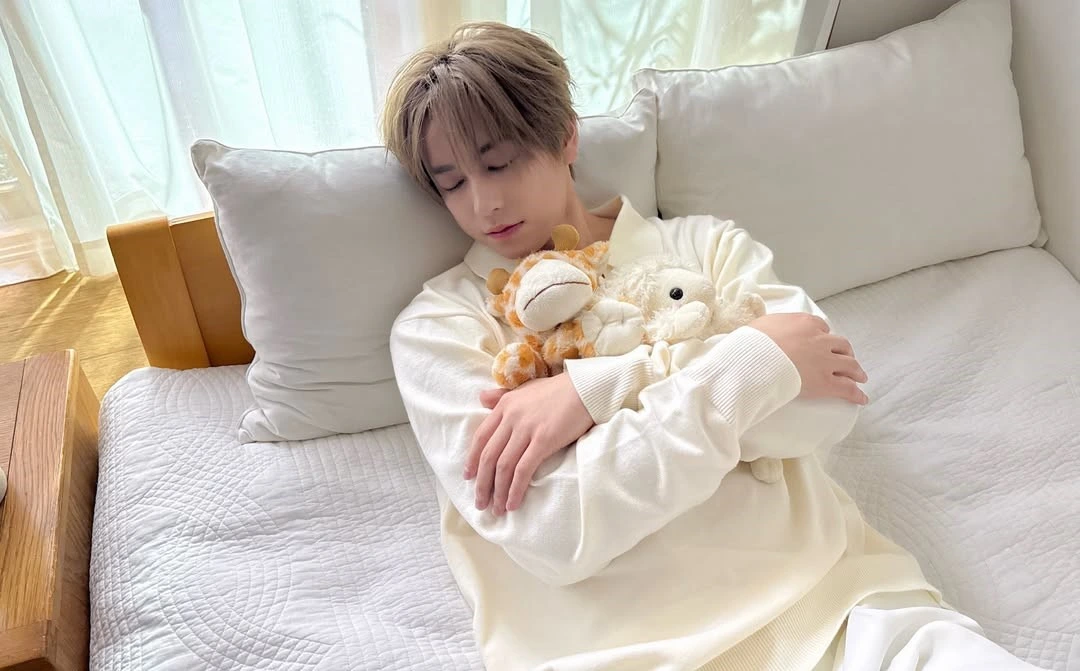 Taki lying on a bed, pretending to be asleep, cuddling two plushies