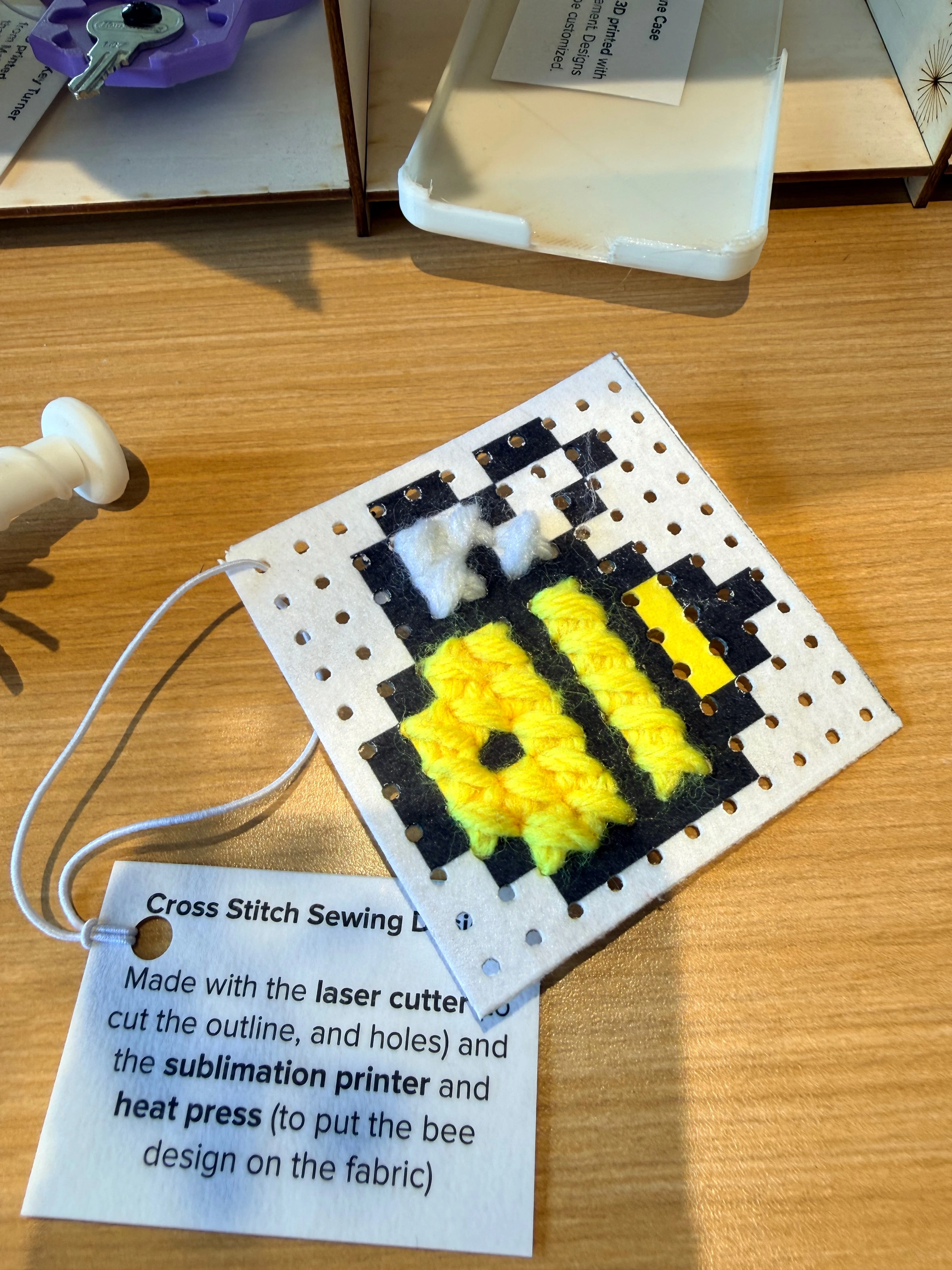 A cross-stitched bee on a small square of fabric. A label tag describing the project is attached to the cross stitch. The label says, "cross stitch sewing.. made with the laser cutter to cut the outline and holes and the sublimation printer and heat press to put the design on the fabric.
