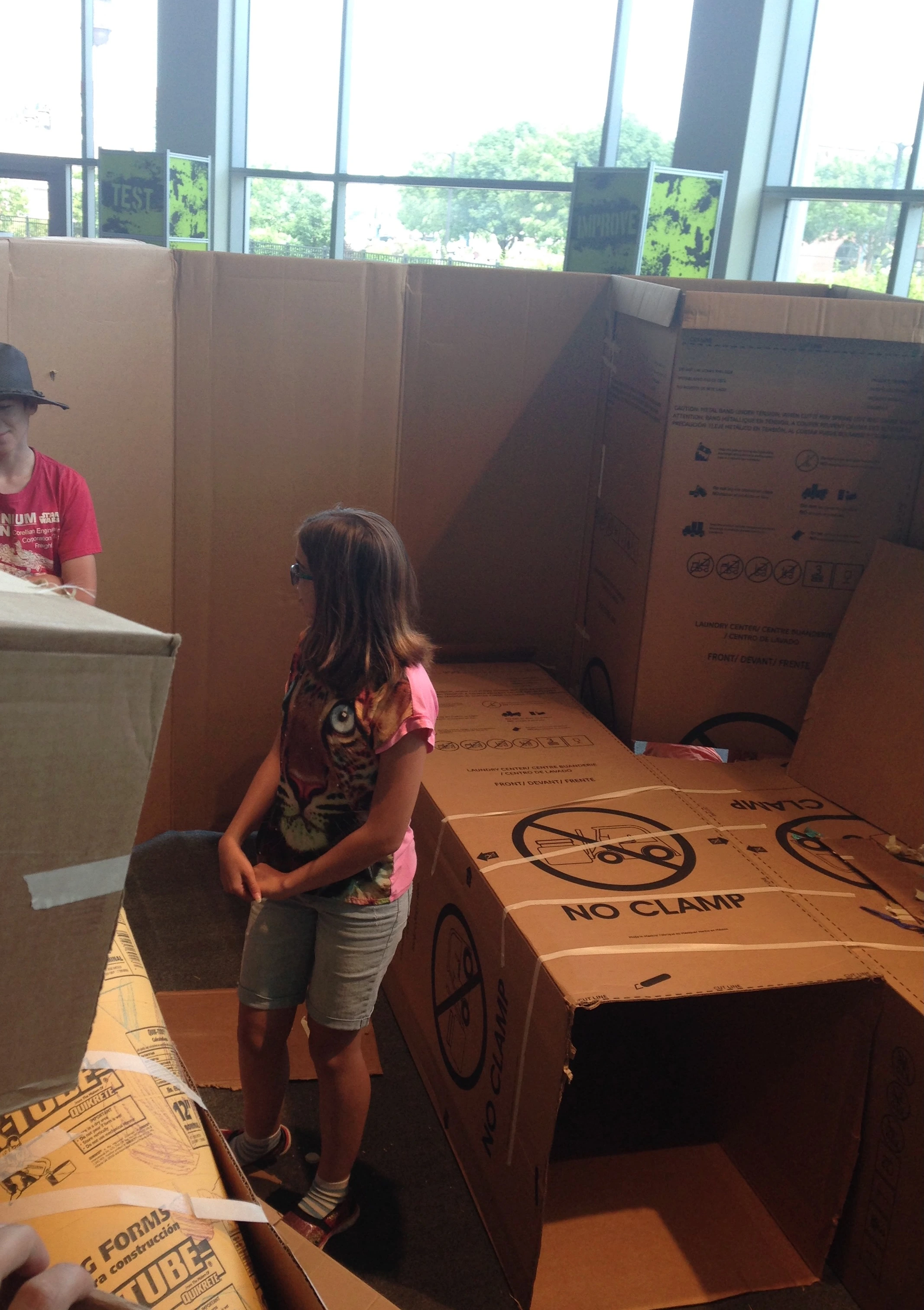 Two young people standing inside of a cardboard construction, large cardboard boxes create walls, tunnels, and tubes that tower over the young people. 