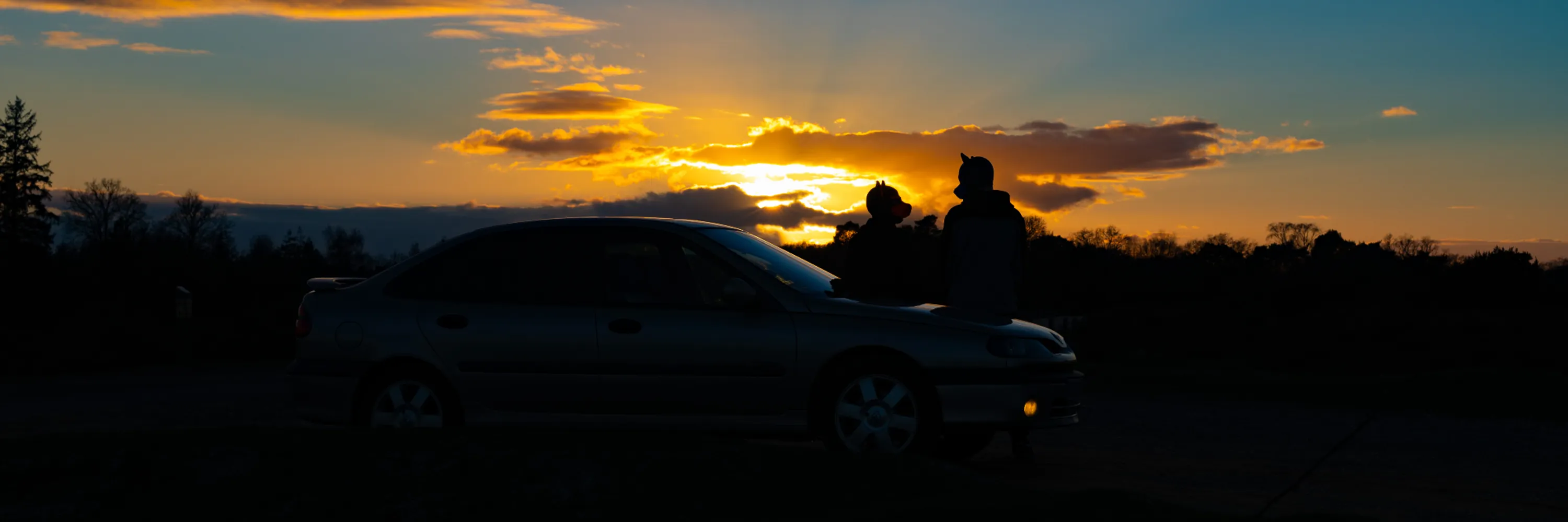 Two human pups sat on the bonnet of a car — a 1999 Renault Laguna 2.9 Monaco V6 — parked up on the edge of a forest, as the sun sets in the background.